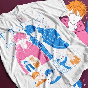 May include: White t-shirt featuring anime-style illustrations of male characters in various poses, with musical notes and Japanese text. The design includes figures in blue, pink, and orange, with a cat graphic.