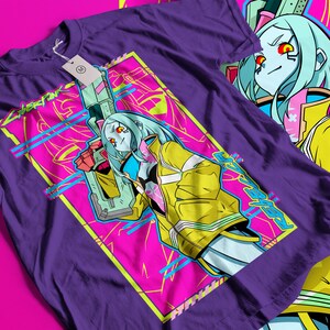 Op de afbeelding: Paars T-shirt met een opvallende grafische afbeelding. Het ontwerp toont een personage met blauw haar, een gele jas en een wapen, tegen een roze en gele achtergrond. Het woord "Cyberpunk" is ook zichtbaar.