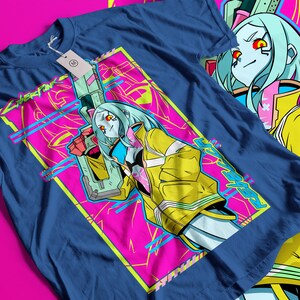 Op de afbeelding: Marineblauw t-shirt met een opvallend grafisch ontwerp. Het ontwerp toont een personage met lichtblauw haar, een gele jas en een wapen, tegen een roze en gele achtergrond. Het woord "CYBERPUNK" is zichtbaar.