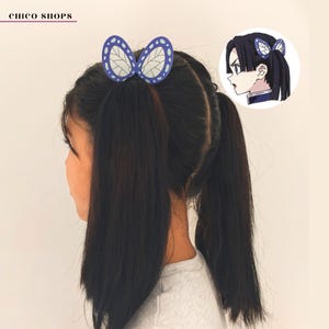 May include: A young person with long, dark hair styled in pigtails, adorned with a blue and white butterfly hair clip. The hair clip is a stylized design, and the image includes a small illustration of the character. The text "chico shops" is in the upper left corner.