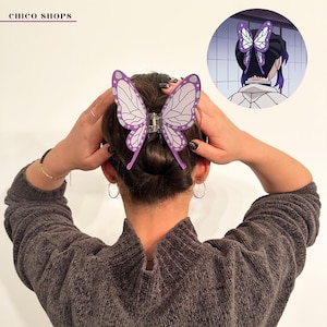 Könnte beinhalten: Eine lila-weiße Haarspange in Schmetterlingsform ist im Haar befestigt. Die Spange ist ein stilisierter Schmetterling mit einem silbernen Verschluss. Die Haarspange ist ein Modeaccessoire. Das Bild enthält auch eine kleine Illustration der Haarspange.