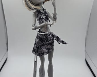 May include: A fashion doll with grey skin, black and white hair, and stitches on the arms and legs. The doll wears a black and white patterned halter top and a matching sarong-style skirt, with grey sandals and a grey plastic display stand.