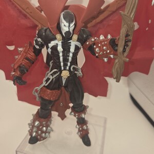 May include: A Spawn action figure featuring a black and white suit, red cape, and spiked gauntlets. The character wears skull-shaped shoulder accents and a belt with a skull buckle. It holds a brown, textured staff and stands on a clear base.