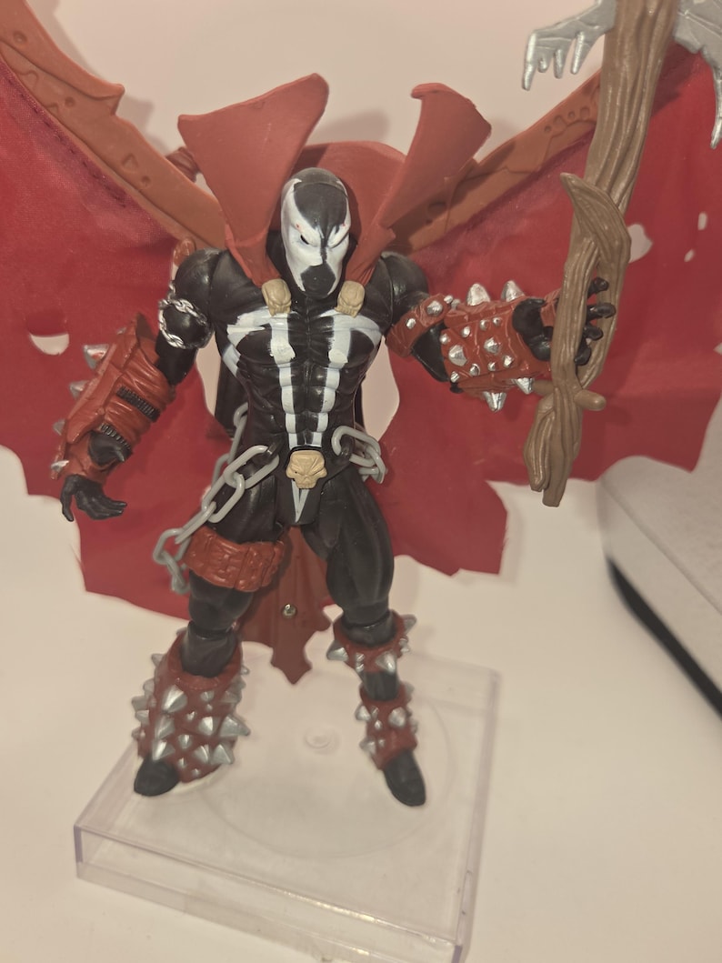 May include: A Spawn action figure featuring a black and white suit, red cape, and spiked gauntlets. The character wears skull-shaped shoulder accents and a belt with a skull buckle. It holds a brown, textured staff and stands on a clear base.