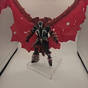 May include: A Spawn action figure featuring a black suit with white chest markings, silver spiked gauntlets and greaves, and a large, tattered red cape with a brown frame. The figure stands on a clear square base with articulated limbs and chains.
