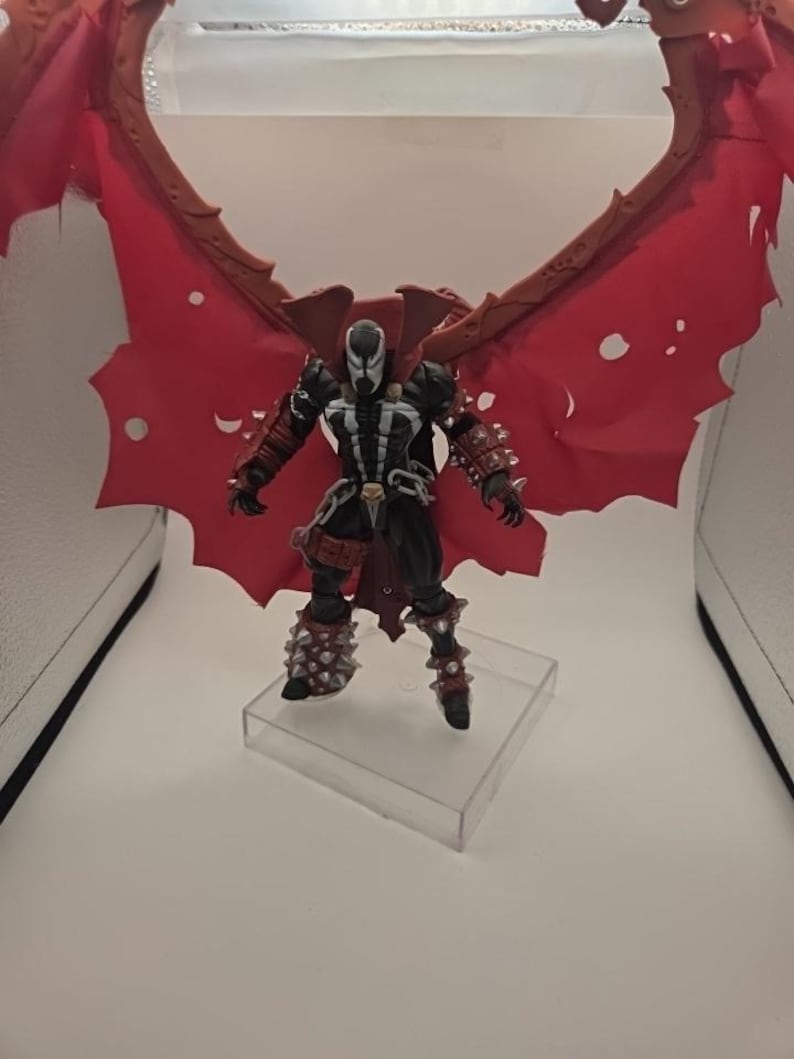 May include: A Spawn action figure featuring a black suit with white chest markings, silver spiked gauntlets and greaves, and a large, tattered red cape with a brown frame. The figure stands on a clear square base with articulated limbs and chains.