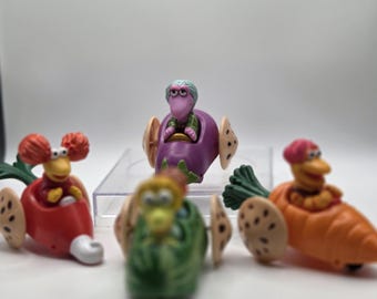 May include: A collection of colorful toy vehicles, each featuring a character. The vehicles are shaped like vegetables, including a red radish, a green watermelon, a purple eggplant, and an orange carrot. The toys have wheels and are set against a white background.