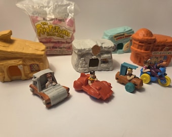 May include: A collection of Flintstones Happy Meal toys, including miniature houses, cars, and figures. The toys are made of colorful plastic and feature characters from the classic cartoon series. A McDonald's Happy Meal bag is in the background.