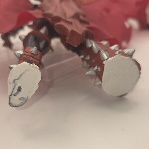 May include: A brown action figure with silver spikes on the legs and feet. The figure features a red cape and white, flat, circular bases attached to the bottom of each foot. The design includes molded details on the legs and a textured surface.
