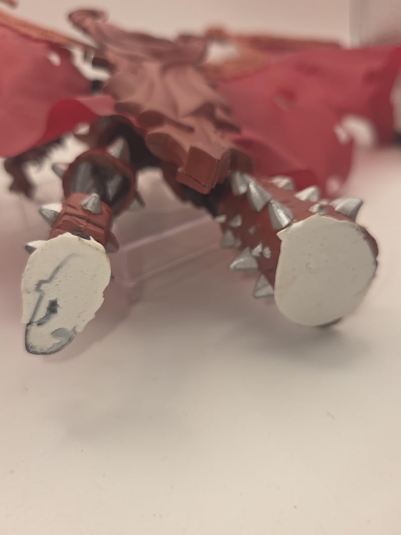 May include: A brown action figure with silver spikes on the legs and feet. The figure features a red cape and white, flat, circular bases attached to the bottom of each foot. The design includes molded details on the legs and a textured surface.