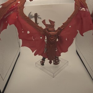 May include: A fantasy action figure featuring large, tattered, reddish-orange wings and a matching hooded cape. The figure has spiked armor on the legs and is mounted on a clear square base, with a grey weapon visible behind the wings.