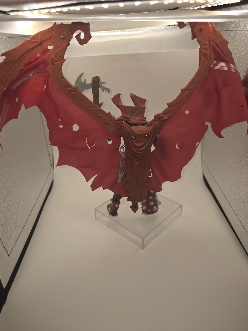 May include: A fantasy action figure featuring large, tattered, reddish-orange wings and a matching hooded cape. The figure has spiked armor on the legs and is mounted on a clear square base, with a grey weapon visible behind the wings.