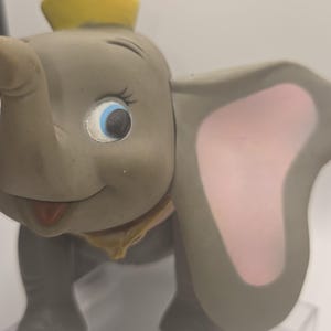 May include: A gray elephant figurine with large ears featuring pink inner sections, blue eyes, and a yellow hat perched on its head. The elephant has a trunk, a small smile, and a yellow collar around its neck, rendered in a smooth, matte finish.