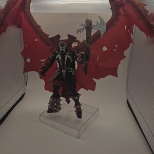 May include: A Spawn action figure featuring a black suit with white accents, a skull belt buckle, and large, tattered red wings. The figure wears spiked gauntlets and boots, and holds a brown, textured weapon with a silver blade, standing on a clear base.