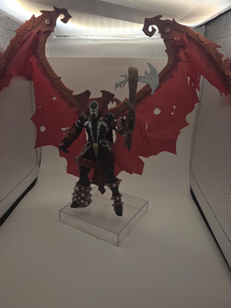 May include: A Spawn action figure featuring a black suit with white accents, a skull belt buckle, and large, tattered red wings. The figure wears spiked gauntlets and boots, and holds a brown, textured weapon with a silver blade, standing on a clear base.