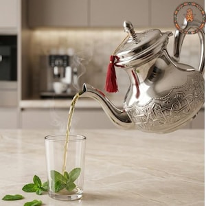 May include: A polished silver teapot with intricate embossed geometric patterns and a red tassel hanging from the lid, pouring steaming tea into a clear glass containing fresh green mint leaves. The teapot features a curved spout and handle.