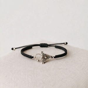 Peut inclure: Bracelet tressé noir avec un charm triangulaire argenté. Le charm présente un motif circulaire au centre et est relié au bracelet par un petit anneau argenté. Le bracelet est réglable grâce à un nœud coulissant.