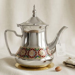 Peut inclure: Une théière en argent poli dotée d'un couvercle pointu avec un fleuron, d'une anse incurvée et d'un long bec. La base est ornée d'un motif floral coloré en rouge, vert et or. Une étiquette blanche indique AGREEABLE HERITAGE, sur un socle doré.