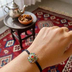 May include: A colorful bracelet with a central, ornate design featuring red, green, and blue accents. The bracelet is on a black cord. A small table with a silver teapot and a bowl of food is in the background.