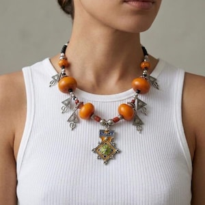 Peut inclure: Un collier artisanal avec de grosses perles orange, des éléments métalliques argentés et un pendentif central avec des détails émaillés colorés. Le collier est porté sur un débardeur blanc côtelé.