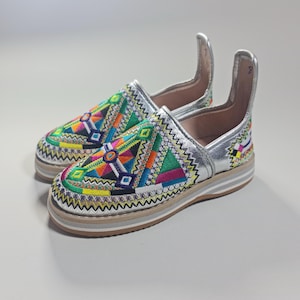 Peut inclure: Une paire de chaussures à enfiler brodées aux couleurs vives. Elles présentent une bordure argentée et une semelle épaisse de couleur claire. La partie supérieure des chaussures est ornée de broderies complexes dans diverses couleurs, dont le vert, le jaune, l'orange et le bleu.