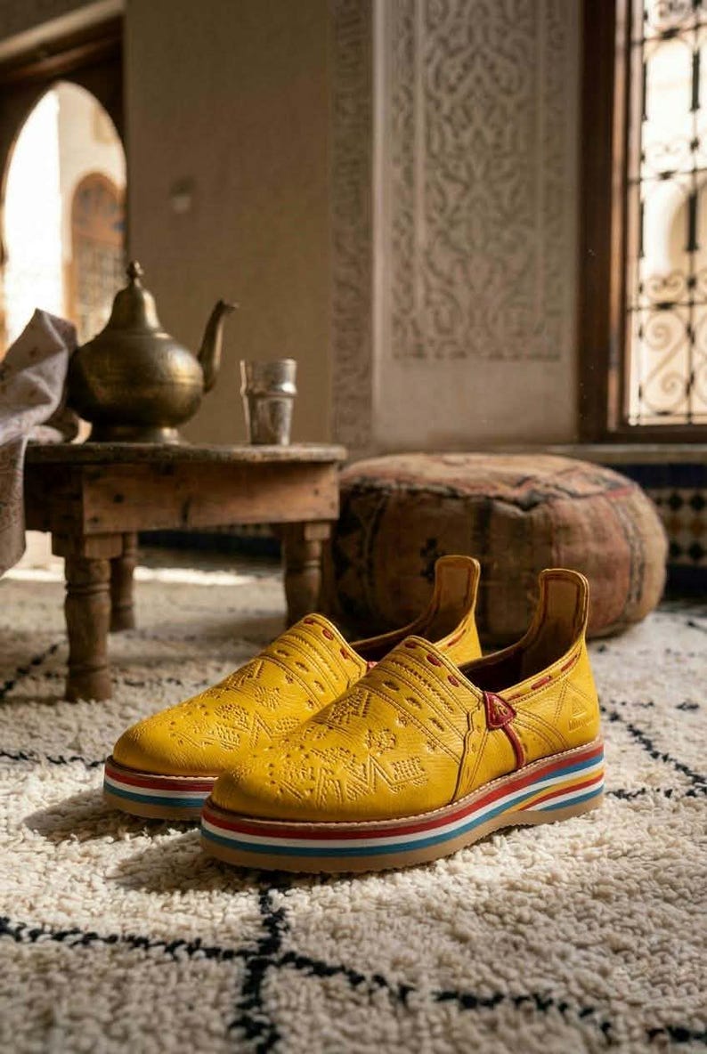 May include: A pair of yellow leather slip-on shoes with intricate embossed patterns. The shoes have a multi-colored striped sole and a high back. They are placed on a white rug with a black geometric design. The background includes a small wooden table and a brass teapot.
