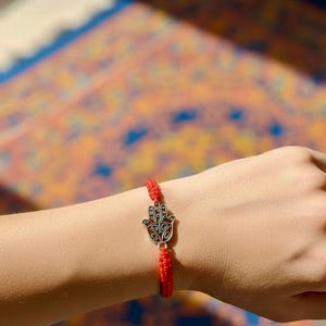 Peut inclure: Un bracelet rouge tressé avec un charm en forme de main de Hamsa de couleur argentée. Le charm est orné de motifs complexes. Le bracelet est porté au poignet, sur un fond coloré à motifs.