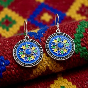 May include: A pair of round, silver-toned earrings with a blue enamel design. The earrings feature a central blue stone surrounded by yellow and green petal-like shapes. The earrings are set against a colorful, patterned textile.