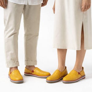 May include: A pair of yellow slip-on shoes with textured leather uppers. The shoes have a red accent on the side and a striped sole. The shoes are worn by two people, with beige pants and a cream-colored dress.