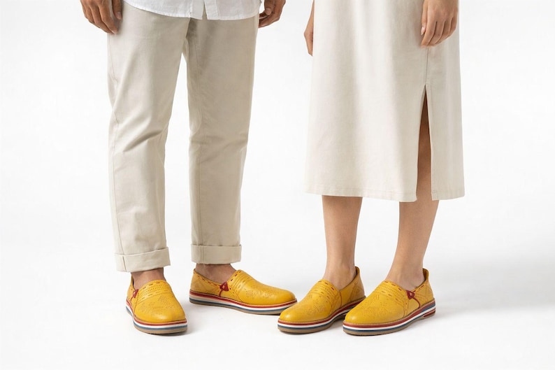 May include: A pair of yellow slip-on shoes with textured leather uppers. The shoes have a red accent on the side and a striped sole. The shoes are worn by two people, with beige pants and a cream-colored dress.