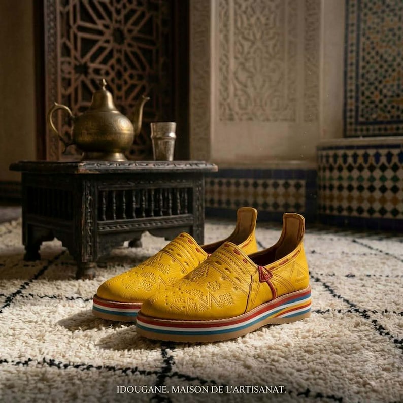 May include: A pair of yellow leather sneakers with intricate embossed patterns. The shoes have a multi-colored striped sole and a red accent on the heel. The text "IDOUGANE. MAISON DE L'ARTISANAT" is at the bottom.