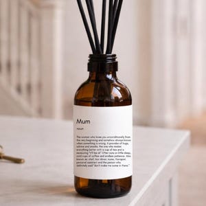 Puede incluir: Un difusor de aromaterapia de vidrio marrón con varillas negras. La botella tiene una etiqueta blanca con la palabra "Mum" y una definición. El difusor está sobre una superficie blanca con llaves y un jarrón de flores en el fondo.