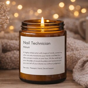 Puede incluir: Una vela encendida en un frasco de vidrio marrón con una etiqueta blanca. La etiqueta dice "Nail Technician" e incluye una definición. La vela está sobre una superficie de madera con una manta suave de color neutro en el fondo.