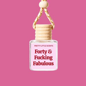 Peut inclure: Un flacon carré en verre pour désodorisant de voiture avec un bouchon en bois clair et une cordelette de suspension. L'étiquette porte le texte PRETTY LITTLE SCENTS et la phrase Forty & Fucking Fabulous en lettres rouges sur fond rose.