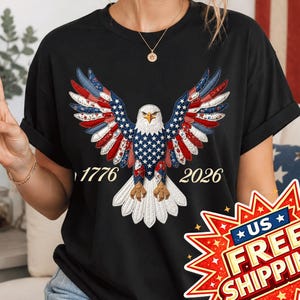 Può includere: T-shirt nero con un design patriottico di aquila. Le ali dell'aquila sono rosse, bianche e blu, e il corpo bianco con un motivo a stelle blu. Le date 1776 e 2026 sono stampate. Maniche corte.