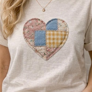 Può includere: T-shirt beige chiaro con un design a cuore patchwork. Il cuore è composto da tessuti floreali, blu e a quadri gialli. È visibile anche una collana d'oro con un piccolo pendente.