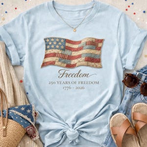 Op de afbeelding: Lichtblauw T-shirt met een verweerd Amerikaans vlagontwerp. De vlag is rood, wit en blauw met een vintage uitstraling. Het woord "Freedom" en de tekst "250 YEARS OF FREEDOM 1776-2026" staan eronder.