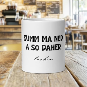 Puede incluir: Una taza de cerámica blanca con el texto "KUMM MA NED A SO DAHER" en letras negras en negrita. La palabra "Enakia" está escrita en cursiva debajo. La taza está sobre una mesa de madera.