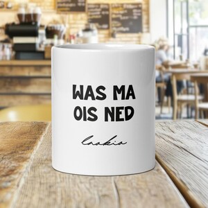 Könnte beinhalten: Eine weiße Keramik-Tasse mit dem Text "WAS MA OIS NED" in schwarzen, fetten Buchstaben. Unter dem Text befindet sich die kursive Unterschrift "Crakia". Die Tasse steht auf einem Holztisch in einem Café.