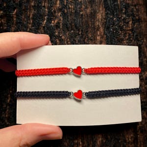 Puede incluir: Dos pulseras de amistad. Una es roja, la otra es negra. Cada pulsera tiene un dije de corazón rojo en el centro. Las pulseras se exhiben en una tarjeta blanca.