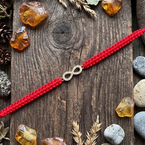 Op de afbeelding: Een rode gevlochten koordarmband met een centraal goudkleurig oneindigheidssymbool ingelegd met kleine heldere steentjes. De armband ligt diagonaal op een donker, verweerd houten oppervlak versierd met barnsteen en dennenappels.