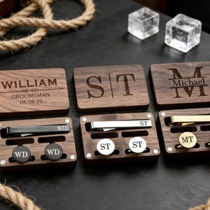 Może przedstawiać: Drewniane zestawy upominkowe z personalizowanymi grawerunkami. Każdy zestaw zawiera spinkę do krawata i spinki do mankietów, zapakowane w drewniane pudełko. Pudełka są grawerowane inicjałami, imionami i datami. Zestawy są prezentowane z kostkami lodu i szklanką bursztynowego płynu.