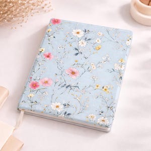 May include: A floral-patterned notebook with a light blue cover. The design features pink, white, and yellow flowers with green stems and leaves. A white ribbon bookmark is visible. The notebook is closed and lies flat on a white surface.