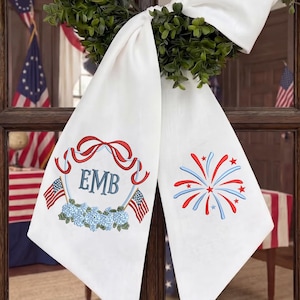 May include: White decorative fabric with embroidered patriotic designs. Features the initials "EMB" within a red ribbon and floral design, American flags, and a firework display in red and blue.