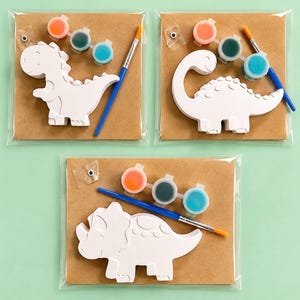 Könnte beinhalten: Drei Bastelsets, die jeweils eine weiße Dinosaurierfigur aus Gips, einen kleinen Pinsel, drei runde Farbtöpfe in Orange, Dunkelgrün und Hellblau sowie ein Wackelauge enthalten, verpackt auf einer braunen Karte in einer transparenten Hülle.