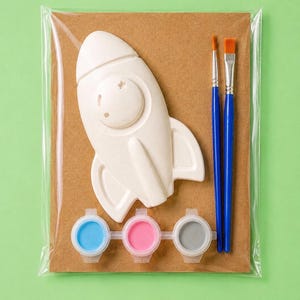 Könnte beinhalten: Eine weiße Raketenfigur aus Keramik, zwei Pinsel mit blauen Griffen und orangefarbenen Borsten sowie drei kleine, runde Farbtöpfe mit hellblauer, rosa und grauer Farbe, alles zusammen verpackt auf einer braunen Pappunterlage.