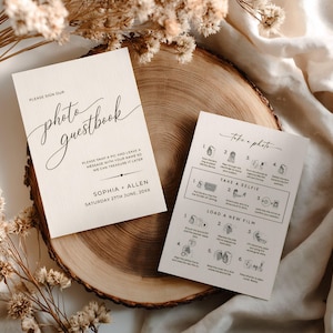 Può includere: Due biglietti color crema con testo nero su una fetta di legno. Un biglietto recita "Photo Guestbook" con un carattere corsivo, e l'altro dice "Take a photo" con istruzioni. Fiori secchi e un panno bianco sullo sfondo.