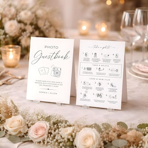 May include: Two white Photo Guestbook signs with black text and illustrations. One sign reads "Photo Guestbook" with instructions and a space for names. The other sign has photo instructions. The signs are on a table with floral arrangements.