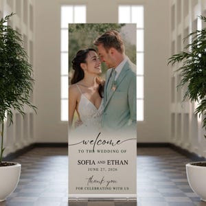 Könnte beinhalten: Ein Hochzeits-Willkommensbanner mit einem Foto eines Paares und dem Text "welcome to the wedding of Sofia and Ethan, June 27, 2026, thank you for celebrating with us." Das Banner wird in einem Flur mit Topfpflanzen angezeigt.