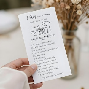 May include: A white card with the text "I Spy" and "photo suggestions" in black script. The card includes a line drawing of a camera and a QR code. The card is held in a hand, with a blurred background.